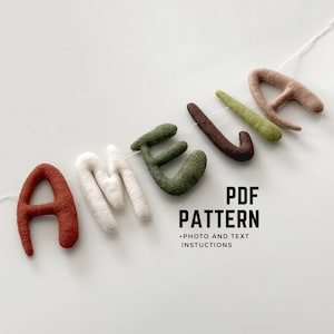 May include: A felt letter banner spelling out the name "Amelia" in various colours, including red, white, brown, green, and beige. The letters are strung together with white thread. The text "PDF PATTERN +PHOTO AND TEXT INSTRUCTIONS" is printed below the banner.