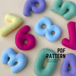May include: A selection of colourful felt numbers in shades of blue, purple, green, pink, and yellow. The numbers are hand-stitched with contrasting thread. The image also includes the text "PDF PATTERN" with "PHOTO AND TEXT INSTRUCTIONS".