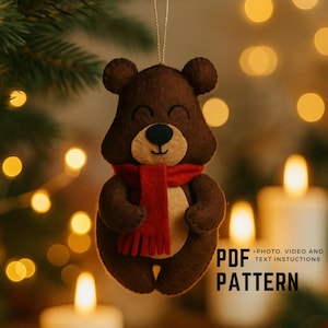 May include: A brown felt bear ornament with a red scarf, black nose, and cream-colored face. The bear has a smiling expression and is suspended by a string. Text on the right side reads "PDF PATTERN".