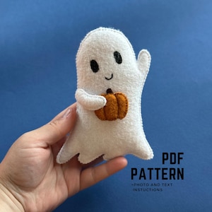 May include: A white felt ghost holding an orange pumpkin. The ghost has black eyes and a smiling mouth. The image includes the text "PDF PATTERN" and "+ PHOTO AND TEXT INSTRUCTIONS" in black font.