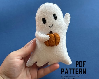 Puede incluir: Un fantasma de fieltro blanco sosteniendo una calabaza naranja. El fantasma tiene ojos negros y una boca sonriente. La imagen incluye el texto "PDF PATTERN" y "+ PHOTO AND TEXT INSTRUCTIONS" en negro.