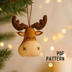 May include: A felt moose ornament with brown antlers, ears, and a smiling face. The moose has a cream-colored muzzle with stitched details. The ornament hangs from a brown string. Text on the right reads "PDF PATTERN +PHOTO AND TEXT INSTUCTIONS."