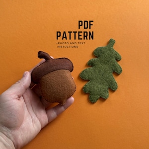 Puede incluir: Una bellota y una hoja de roble de fieltro sobre un fondo naranja. La bellota es marrón con una tapa marrón oscuro. La hoja es verde. El texto en la imagen dice "PDF PATTERN + PHOTO AND TEXT INSTRUCTIONS".