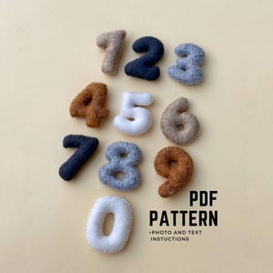 May include: A selection of felt numbers in diverse colours, such as brown, navy, and white, set against a beige backdrop. The numbers range from zero to nine. The text "PDF PATTERN + PHOTO AND TEXT INSTRUCTIONS" is visible.