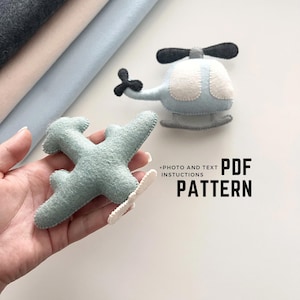 May include: A felt airplane and helicopter in shades of blue and white. The airplane is held in a hand. The text "PDF PATTERN" is displayed in black. The image also includes the text "+PHOTO AND TEXT INSTRUCTIONS".