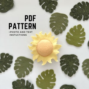May include: A yellow felt sun with a yellow center and a yellow felt border. The sun is surrounded by green felt monstera leaves. The text "PDF PATTERN +PHOTO AND TEXT INSTRUCTIONS" is written in black text on a white background.