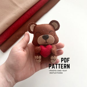 May include: A brown felt bear holding a red felt heart. The bear has a light brown muzzle and ears. The image also shows a stack of felt in shades of brown and red. Text on the image reads "PDF PATTERN + PHOTO AND TEXT INSTRUCTIONS."