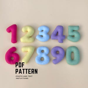 May include: A set of plush numbers in various colours, including pink, yellow, light blue, purple, and green. The numbers from zero to nine are displayed. The text "PDF PATTERN" and "+PHOTO AND TEXT INSTRUCTIONS" are below the numbers.