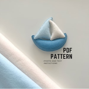 Pode incluir: Um padrão PDF para um brinquedo de veleiro de feltro. O padrão inclui instruções e fotos. A imagem mostra um veleiro de feltro azul e branco sobre um fundo branco.