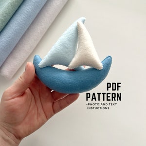 May include: A blue felt sailboat with a white sail. The sailboat is being held in a hand. The text "PDF PATTERN +PHOTO AND TEXT INSTRUCTIONS" is below the sailboat.