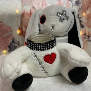 Puede incluir: Conejo de peluche blanco con orejas negras y un corazón rojo en el pecho. El conejo lleva un collar negro con tachuelas y tiene un tarro de vidrio transparente dentro de su cuerpo.