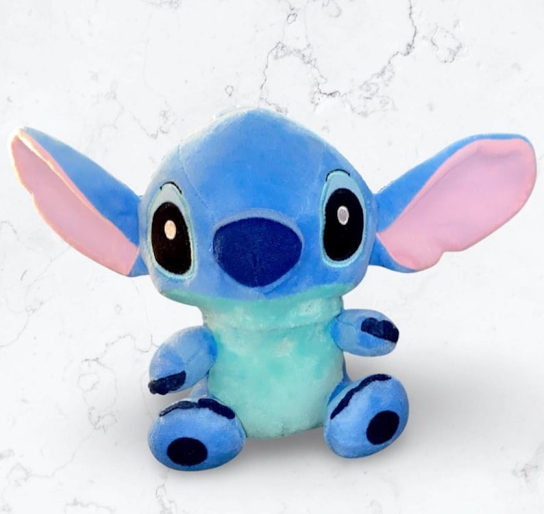 May include: A blue and pink plush toy of Stitch from the Disney movie Lilo & Stitch. The toy is sitting with its legs crossed and has a big, friendly smile.