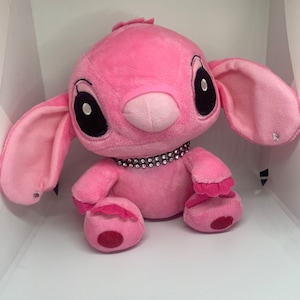 May include: Pink plush toy with large ears, black eyes, and a sparkly pink collar. The toy is sitting on a white surface.