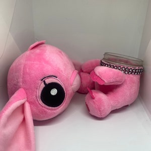 May include: Two pink plush toys, one with large eyes and floppy ears, the other holding a clear glass jar with a pink rhinestone trim.