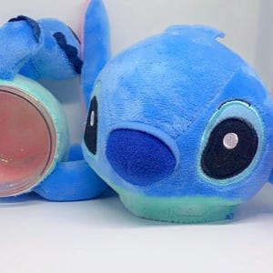 May include: Blue plush toy of a cartoon character with large eyes, a pink nose, and pink ears. The toy is holding a clear jar with pink glitter inside.