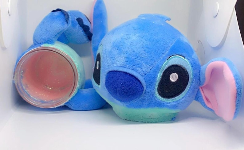 May include: Blue plush toy of a cartoon character with large eyes, a pink nose, and pink ears. The toy is holding a clear jar with pink glitter inside.