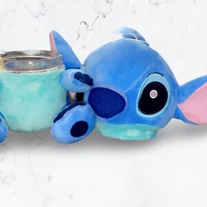 May include: Blue Stitch plush toy holding a clear glass with a blue and green interior. The Stitch plush has a pink ear and a black eye.