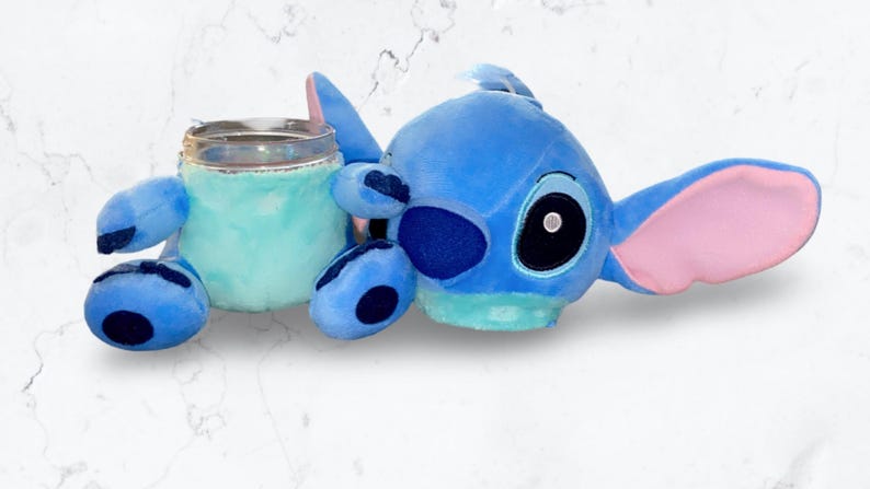 May include: Blue Stitch plush toy holding a clear glass with a blue and green interior. The Stitch plush has a pink ear and a black eye.