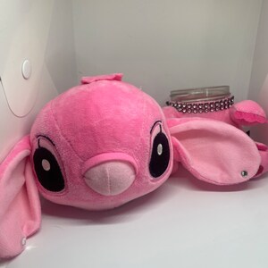 May include: Pink plush toy shaped like a cartoon character with large black eyes and a small pink nose. The toy is sitting on a white surface next to a clear glass jar with a sparkly pink lid.
