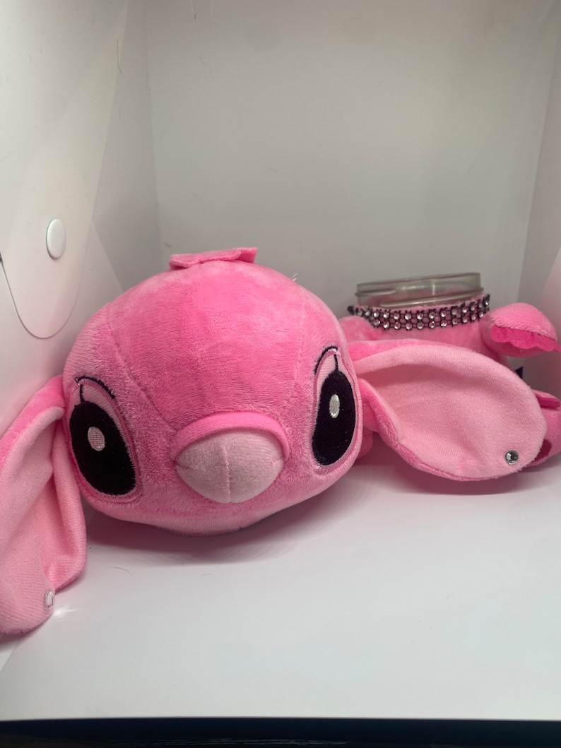 May include: Pink plush toy shaped like a cartoon character with large black eyes and a small pink nose. The toy is sitting on a white surface next to a clear glass jar with a sparkly pink lid.