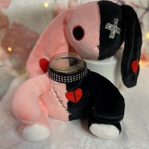 May include: A pink and black plush bunny with a broken heart patch and a studded black band around its neck. The bunny has a black eye and a white cross with rhinestones on its head.