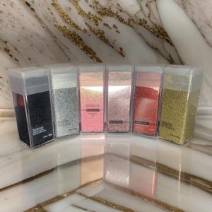 May include: Seven small clear plastic containers filled with different colored glitter. The containers are labeled with the word "Beautifullover" and the color of the glitter. The colors are black, silver, pink, rose gold, red, and gold.