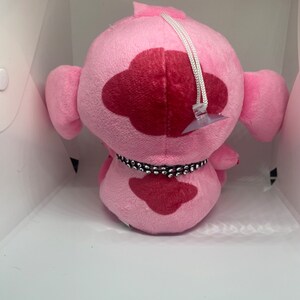 May include: Pink plush toy with a red heart-shaped patch and a black beaded collar. The toy has a white string attached to the top of its head.