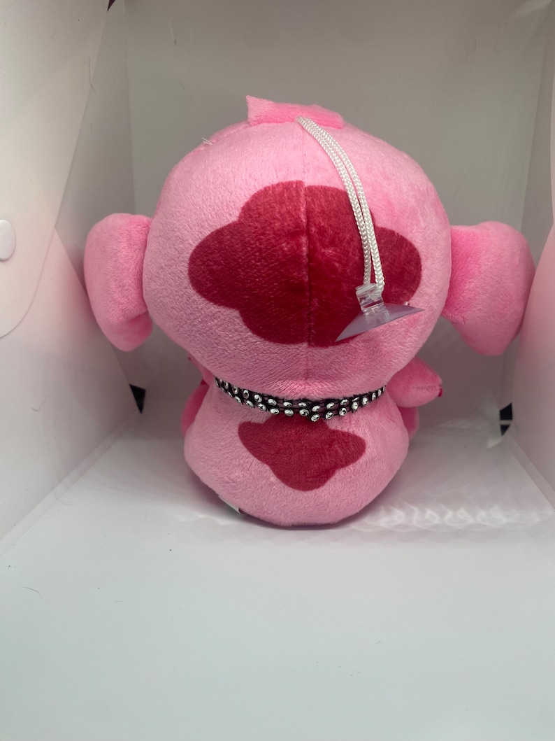 May include: Pink plush toy with a red heart-shaped patch and a black beaded collar. The toy has a white string attached to the top of its head.