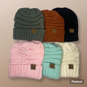 May include: Six knitted beanies in various colors, including green, orange, black, pink, light blue, and cream. Each beanie has a small brown leather tag with the letters "CC" on it.
