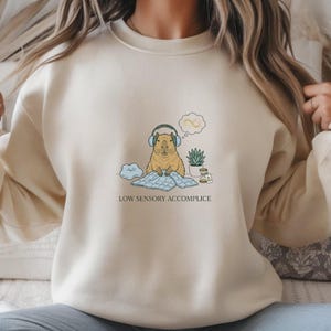 Puede incluir: Sudadera color crema con un gráfico de un carpincho con auriculares, sentado sobre una manta azul, y una burbuja de pensamiento con un símbolo de infinito. El texto "LOW SENSORY ACCOMPLICE" está impreso debajo.