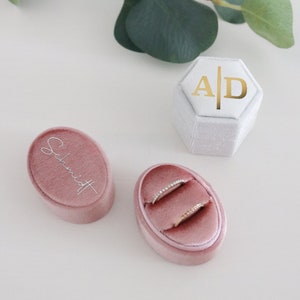 May include: A collection of ring boxes in a soft, romantic setting. Two pink velvet oval ring boxes and a white hexagonal box with gold lettering. The oval boxes have the name "Schmidt" and rings inside. Eucalyptus leaves are in the background.