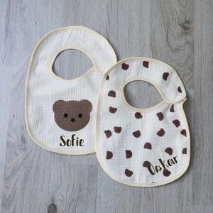 May include: Two white cotton baby bibs with brown bear designs. One bib has a brown bear face and the name "Sofie" embroidered on it. The other bib has a brown bear print pattern and the name "Oskar" embroidered on it.