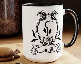 May include: White ceramic mug with a black rim and handle. The mug features a black and white illustration of a druid symbol with two dragons, a staff, and dice. The text "DRUID" is printed on the mug.