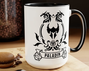 May include: White ceramic mug with a black rim and handle. The mug features a black and white design of two dragons flanking a shield with a dice and the word "PALADIN" on a banner.