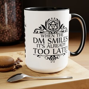 May include: White ceramic mug with a black rim and handle. The mug features a black and white illustration of a 20-sided die with a decorative border and the text "When the DM smiles it's already too late!"