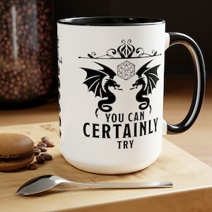 May include: White ceramic mug with a black rim and handle. The mug features a black and white illustration of two dragons flanking a twenty-sided die. The text "You can certainly try" is printed below the illustration.