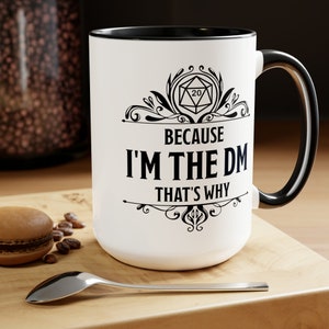 May include: White ceramic mug with a black rim and handle. The mug features a black and white design of a 20-sided die with a decorative border and the text "Because I'm the DM That's Why".