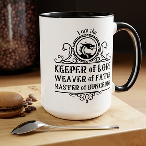 May include: White ceramic mug with a black rim and handle. The mug features a black and white design with a dragon, the text "I am the Keeper of Lore, Weaver of Fates, Master of Dungeons", and decorative flourishes.