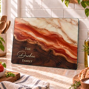 May include: A rectangular glass cutting board featuring a marbled design with cream, deep red, and dark brown wood-grain patterns. The text "THE Durkee FAMILY" is printed in white on the lower left side of the board, which is positioned upright.