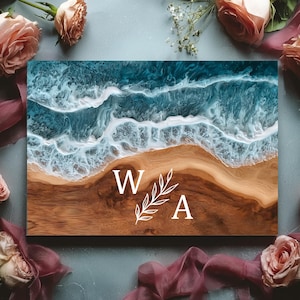 May include: A wooden sign with a resin ocean design. The top half shows blue and white waves, leading to a brown beach. White letters "W A" and a leaf design are in the center. Pink roses and burgundy fabric surround the sign.