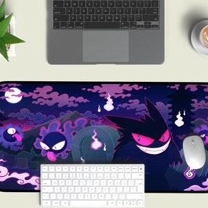 May include: A black and purple gaming mouse pad with a design of a cartoon ghost with glowing pink eyes. The ghost is surrounded by other smaller ghosts and clouds. The mouse pad is on a white surface with a keyboard and a coffee cup.