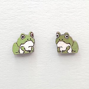 May include: A pair of stud earrings featuring a flat, illustrated frog design. Each frog is colored in shades of light green with a white belly, black outlines, and large white eyes with black pupils, rendered in a simple, graphic style.