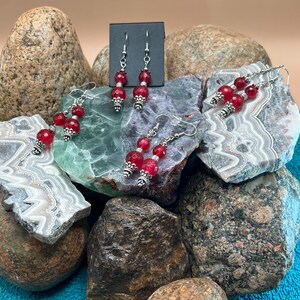 May include: Red beaded dangle earrings with silver-colored accents. The earrings feature faceted red beads and decorative silver-toned metal components. The earrings are displayed on a variety of rocks.