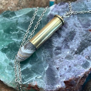 May include: A silver chain necklace featuring a pendant made from a brass bullet casing and a polished stone. The stone has a mix of brown, gray, and white patterns. The necklace is displayed on a green and purple stone.