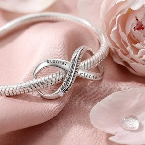May include: A silver snake chain bracelet featuring an infinity symbol charm. The charm is engraved with the word eternal on both sides and includes a small clear stone accent. The metal surface has a polished finish with detailed texture.