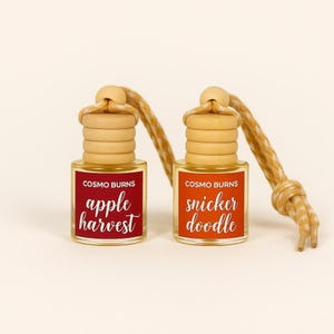 Könnte beinhalten: Zwei kleine Glasflaschen mit Holzaufsätzen und Aufhängeschnüren. Eine Flasche hat ein rotes Etikett mit dem Text "apple harvest", die andere ein orangefarbenes Etikett mit dem Text "snicker doodle". Die Flaschen sind für Duftstoffe.