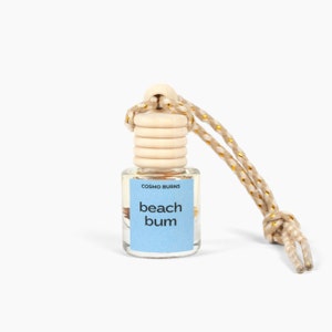 Peut inclure: Petite bouteille en verre transparent avec un bouchon en bois et une corde beige. L'étiquette bleu clair porte les mots "beach bum" et "Cosmo Burns". La corde est ornée de fils dorés.