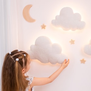 May include: A wall-mounted lighting set featuring two white, textured, cloud-shaped lamps that emit a soft glow. The arrangement includes a smooth, light-coloured wooden crescent moon and three small wooden star accents mounted on the wall.