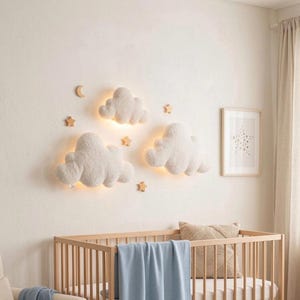 Peut inclure: Intérieur de chambre d'enfant avec un lit bébé en bois clair et une couverture bleue. Au-dessus du lit, trois lampes murales en forme de nuage et des décorations en forme d'étoiles et de lune en bois sont fixées au mur.