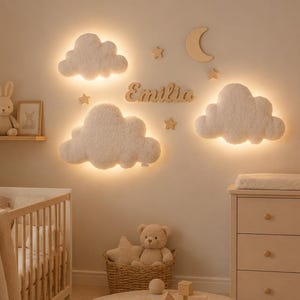May include: Three white, fluffy, cloud-shaped wall lights with warm backlighting, arranged with wooden star and moon cutouts and a central wooden name sign reading Emilio. The wall decor is displayed above a wooden crib and a dresser.